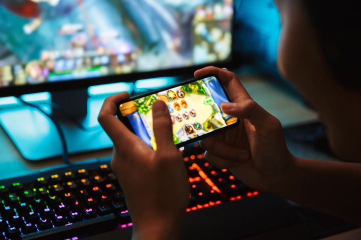 Portrait of teenage gamer boy playing video games on smartphone and computer in dark room wearing headphones and using backlit colorful keyboard By Dean Drobot | www.shutterstock.com