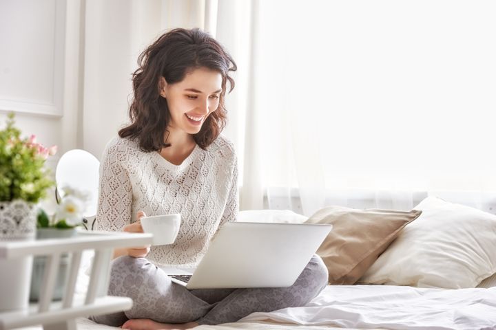 Woman working on a laptop sitting on the bed in the house. By Yuganov Konstantin | www.shutterstock.com